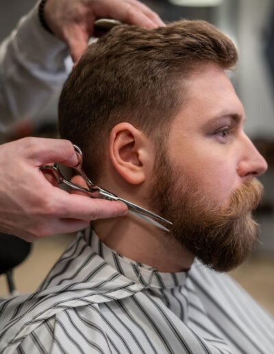 A side view of a barber trimming a bearded man's hair in a barber shop.